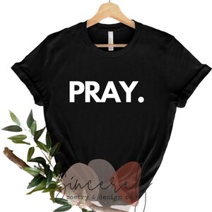 May include: Black t-shirt with the word "PRAY." in bold white letters. The shirt is displayed on a wooden hanger. The bottom of the image features a decorative arrangement of leaves and a branch, along with the text "Sincere poetry & design co."