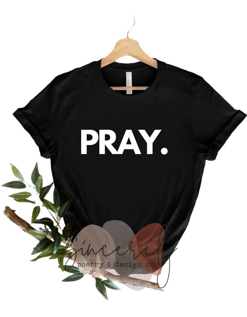 May include: Black t-shirt with the word "PRAY." in bold white letters. The shirt is displayed on a wooden hanger. The bottom of the image features a decorative arrangement of leaves and a branch, along with the text "Sincere poetry & design co."