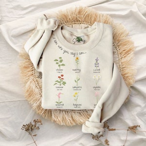 May include: A cream-coloured sweatshirt featuring floral illustrations and the phrase "I am who you say I am...". The design incorporates various flowers with words such as "chosen", "worthy", and "loved". The sweatshirt is folded on a woven mat.