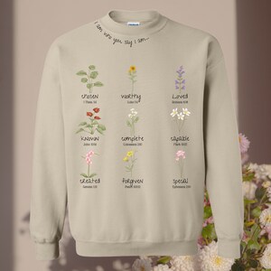 May include: Beige sweatshirt featuring floral designs and the words "chosen", "worthy", "loved", "known", "complete", "capable", "created", "forgiven", and "special". The neckline reads "I am who you say I am..."