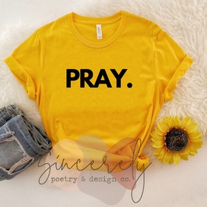 May include: Mustard yellow t-shirt with the word "PRAY." in black block letters. Rolled sleeves and a crew neck. The shirt is paired with blue denim jeans and a sunflower. The words "Sincerely poetry & design co." are in the bottom left corner.