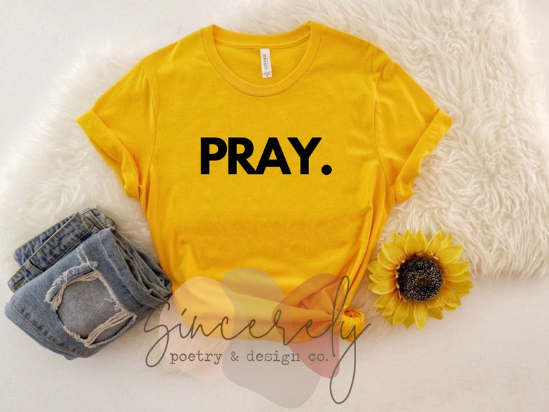 May include: Mustard yellow t-shirt with the word "PRAY." in black block letters. Rolled sleeves and a crew neck. The shirt is paired with blue denim jeans and a sunflower. The words "Sincerely poetry & design co." are in the bottom left corner.
