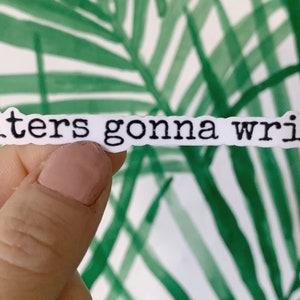 May include: A white sticker with the phrase "Writers gonna write" in black, typewriter-style font. The sticker is held against a background of green and white tropical leaves.