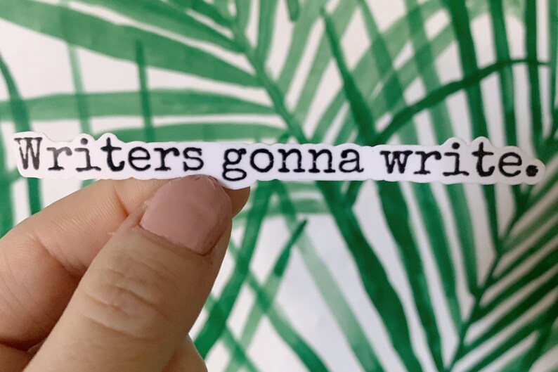May include: A white sticker with the phrase "Writers gonna write" in black, typewriter-style font. The sticker is held against a background of green and white tropical leaves.