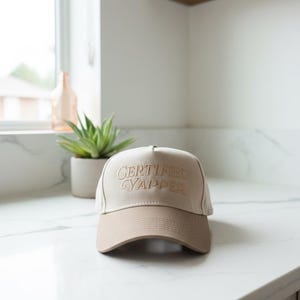 Peut inclure: Casquette de baseball beige et marron avec le texte brodé "CERTIFIED YAPPER". Le chapeau est posé sur un comptoir blanc. Une petite plante en pot et une fenêtre sont en arrière-plan.