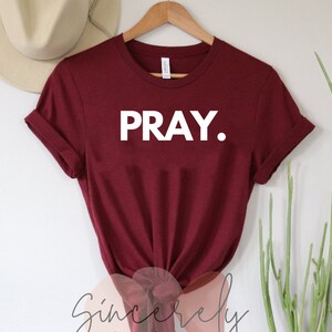 May include: A maroon t-shirt with the word "PRAY." in bold white letters. The shirt is hanging on a wooden hanger. The brand name "Sincerely poetry & design co." is visible at the bottom of the shirt.