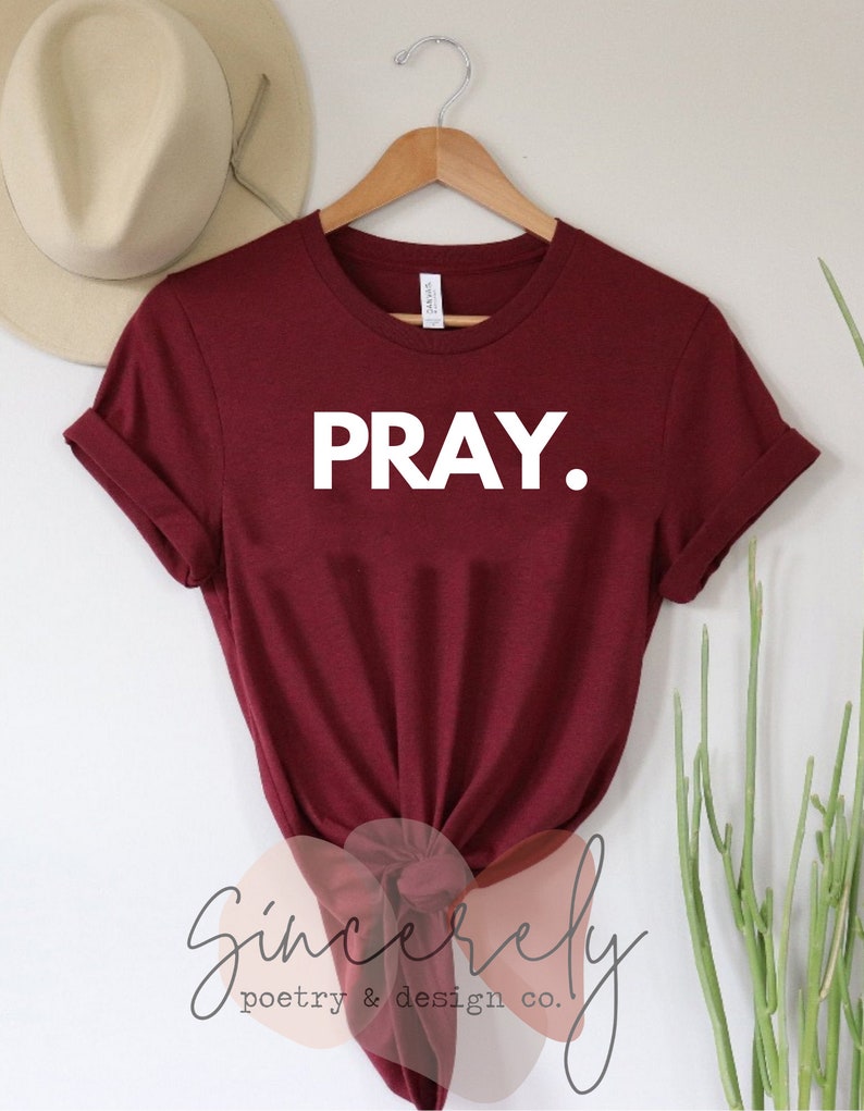 May include: A maroon t-shirt with the word "PRAY." in bold white letters. The shirt is hanging on a wooden hanger. The brand name "Sincerely poetry & design co." is visible at the bottom of the shirt.
