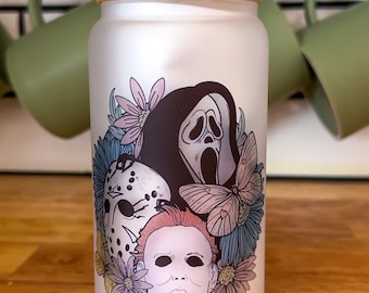 May include: Frosted glass can-shaped tumbler with a bamboo lid. The design features horror movie characters, including Ghostface, Jason Voorhees, and Michael Myers, surrounded by floral and butterfly accents. A unique drinkware item.
