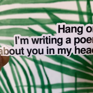 May include: A white sticker with black text that reads "Hang on. I'm writing a poem about you in my head." The sticker is held up against a background of green and white palm leaves.