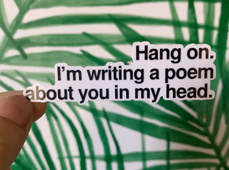 May include: A white sticker with black text that reads "Hang on. I'm writing a poem about you in my head." The sticker is held up against a background of green and white palm leaves.
