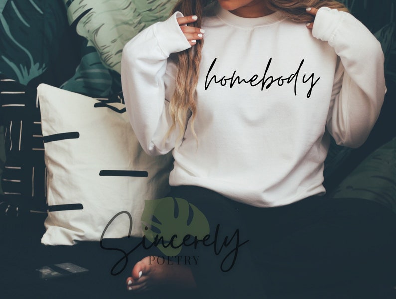 May include: A white sweatshirt with the word "homebody" in black script. The sweatshirt is long-sleeved and has a crew neck. The image also shows a decorative pillow and a leafy background.