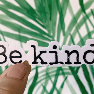May include: A white sticker with the words "Be kind." in black, bold letters. The sticker is held in front of a green and white tropical leaf background.
