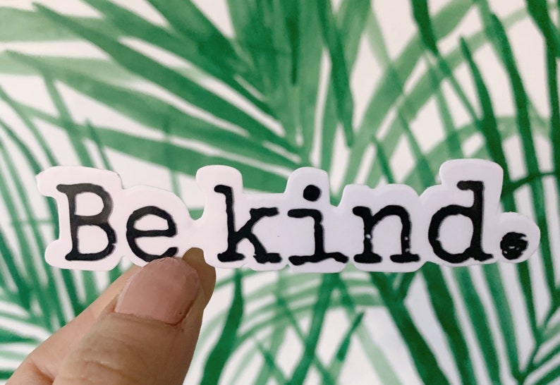 May include: A white sticker with the words "Be kind." in black, bold letters. The sticker is held in front of a green and white tropical leaf background.