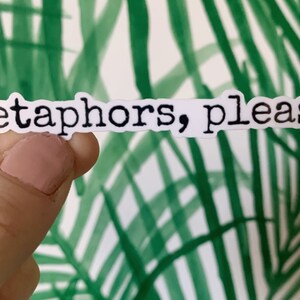 May include: A white sticker with the phrase "Metaphors, please." in black, typewriter-style font. The sticker is held against a background of green and white palm frond design.