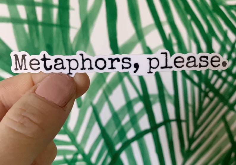 May include: A white sticker with the phrase "Metaphors, please." in black, typewriter-style font. The sticker is held against a background of green and white palm frond design.
