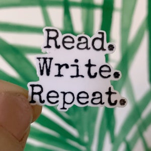 May include: A white sticker with the words "Read. Write. Repeat." in black, stacked vertically. The sticker is held against a background of green and white abstract shapes.