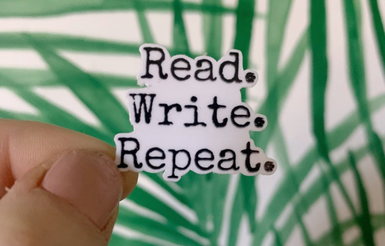 May include: A white sticker with the words "Read. Write. Repeat." in black, stacked vertically. The sticker is held against a background of green and white abstract shapes.