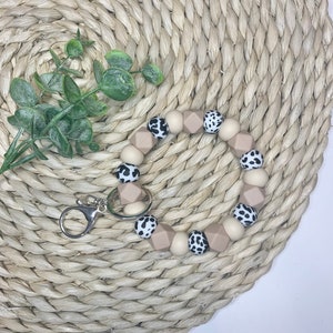 May include: A beige and black silicone keychain with a cow print design. The keychain is made of round and hexagonal beads and has a silver metal clasp.