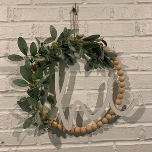 May include: A round wooden wreath with the word "home" cut out in white. The wreath is decorated with faux greenery and wooden beads. It hangs against a white brick wall.