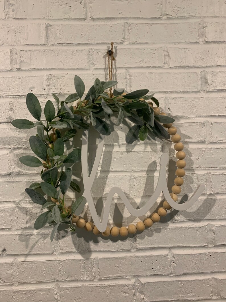 May include: A round wooden wreath with the word "home" cut out in white. The wreath is decorated with faux greenery and wooden beads. It hangs against a white brick wall.
