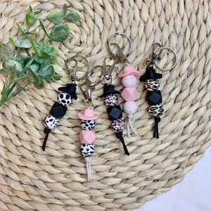 May include: Four keychains with black and white cow print, leopard print, and pink and white cow print beads. Each keychain has a small black cowboy hat charm and a pink cowboy hat charm.
