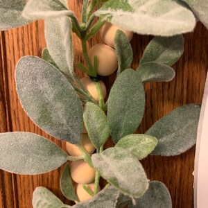May include: Close-up of a decorative garland featuring green leaves with a soft, velvety texture and small, round, light-colored wooden beads. The garland is set against a wooden background, creating a natural and rustic aesthetic.