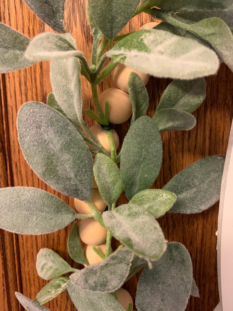 May include: Close-up of a decorative garland featuring green leaves with a soft, velvety texture and small, round, light-colored wooden beads. The garland is set against a wooden background, creating a natural and rustic aesthetic.