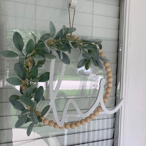 May include: A round wreath with the word "Welcome" in white script. The wreath is decorated with faux green leaves, wooden beads, and is hanging on a door. The wreath is suspended by a white over-the-door hanger.
