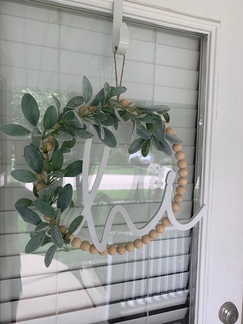May include: A round wreath with the word "Welcome" in white script. The wreath is decorated with faux green leaves, wooden beads, and is hanging on a door. The wreath is suspended by a white over-the-door hanger.