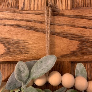May include: A close-up of a decorative wreath with wooden beads and faux greenery. The wreath is suspended by a jute rope from a metal hook. The background is a wooden door with a natural wood grain.