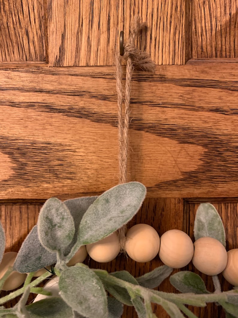 May include: A close-up of a decorative wreath with wooden beads and faux greenery. The wreath is suspended by a jute rope from a metal hook. The background is a wooden door with a natural wood grain.