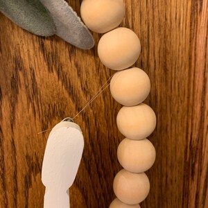 May include: Close-up of a wooden bead garland with a white decorative element and a faux green leaf. The beads are light brown and round, strung together with a thin wire. The background is a dark wood surface.