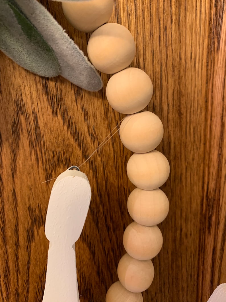May include: Close-up of a wooden bead garland with a white decorative element and a faux green leaf. The beads are light brown and round, strung together with a thin wire. The background is a dark wood surface.