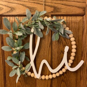 May include: A round wooden wreath with the word "Hi" in white script. The wreath is adorned with light green faux leaves and small wooden beads. The wreath hangs from a natural twine string.