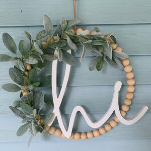 May include: A white wooden "Home" sign with a wreath design. The wreath features faux sage leaves, wooden beads, and a jute string for hanging. The sign is displayed against a light blue background.