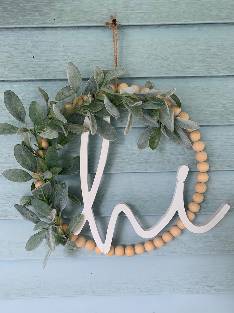 May include: A white wooden "Home" sign with a wreath design. The wreath features faux sage leaves, wooden beads, and a jute string for hanging. The sign is displayed against a light blue background.