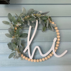 May include: A round, white wooden sign with the word "hi" in a cursive font. The sign is decorated with faux green leaves and small, light-colored wooden beads. The sign is hanging on a light blue wooden wall.