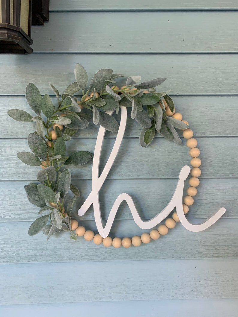 May include: A round, white wooden sign with the word "hi" in a cursive font. The sign is decorated with faux green leaves and small, light-colored wooden beads. The sign is hanging on a light blue wooden wall.