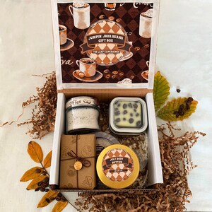 May include: A gift box labeled "Jumpin Java Beans" filled with coffee-themed items. The box contains a candle, soap, body butter, and a bar of soap. The box is lined with brown paper and coffee bean accents. The lid features coffee cups.