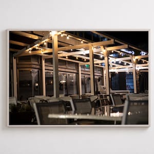 May include: A framed wall art print depicting an outdoor restaurant patio at night. The scene features a wooden pergola structure illuminated by strings of warm glowing Edison bulbs, with tables and chairs arranged underneath on a dark terrace.
