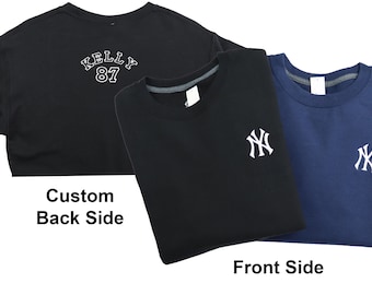 Op de afbeelding: Twee zwarte sweatshirts met witte borduursels. Eén sweatshirt heeft "KELLY 87" op de rug. Het andere sweatshirt heeft een New York Yankees logo op de voorkant. Een blauw sweatshirt met een wit New York Yankees logo ligt op het zwarte sweatshirt.