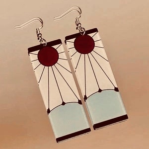May include: A pair of silver earrings with white and red rectangular charms. The charms feature a sun with rays and a red circle in the center.