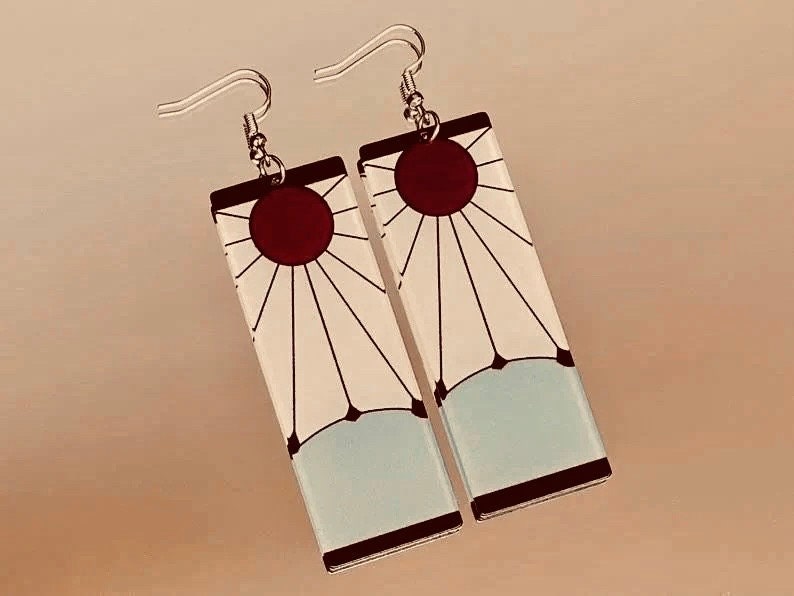 May include: A pair of silver earrings with white and red rectangular charms. The charms feature a sun with rays and a red circle in the center.