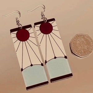 May include: A pair of earrings featuring a white and blue design with a red sun in the center. The earrings are rectangular and have a silver hook.