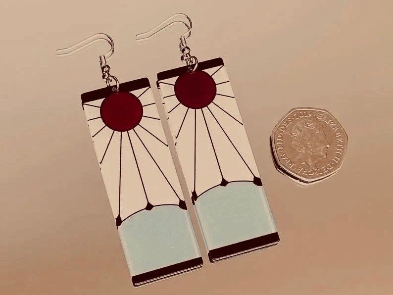May include: A pair of earrings featuring a white and blue design with a red sun in the center. The earrings are rectangular and have a silver hook.
