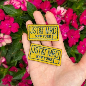 May include: Two rectangular embroidered patches featuring a yellow background with a dark blue border and dark blue text that reads JST&T MRD NEW YORK. The patches have a stitched finish and are rectangular in shape with rounded corners.