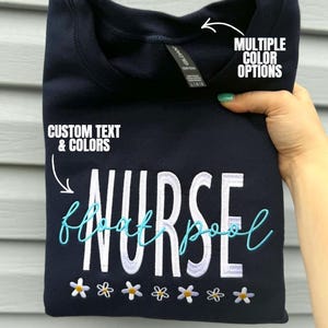 May include: Navy blue sweatshirt with the word "NURSE" in white block letters and "float pool" in turquoise script. The design includes small embroidered flowers. The text "CUSTOM TEXT & COLORS" and "MULTIPLE COLOR OPTIONS" are also visible.