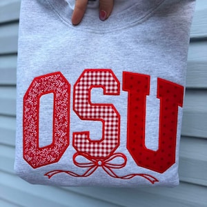 Peut inclure: Un sweat-shirt gris clair à col rond avec les lettres OSU en appliqué rouge. Le O présente un motif floral blanc, le S un motif à carreaux vichy rouge et blanc, et le U un motif à pois rouges. Un nœud brodé rouge est situé sous le S.