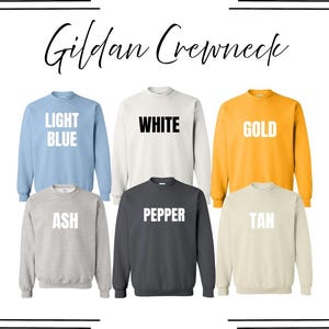 May include: A collection of six Gildan Crewneck sweatshirts displayed in a grid. The colors include light blue, white, gold, ash, pepper, and tan. Each sweatshirt features its respective color name printed in bold text on the front of the garment.