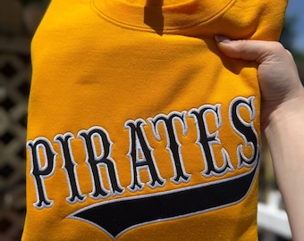 May include: A bright yellow crewneck sweatshirt featuring the word PIRATES embroidered in black lettering with a white outline. The text is positioned above a black swooping underline detail that is also outlined in white thread.
